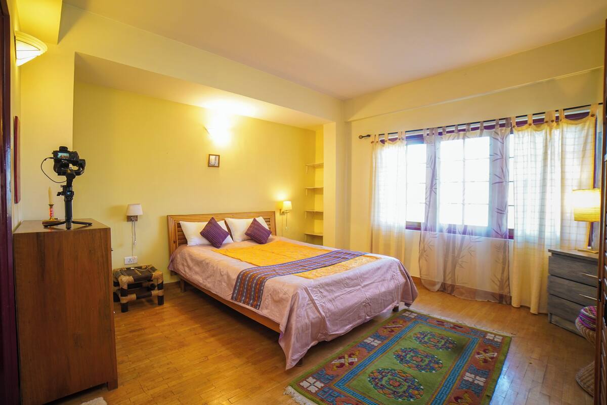 A bedroom with a bed, wooden floor, a desk with a camera, a lamp, and a window with curtains.