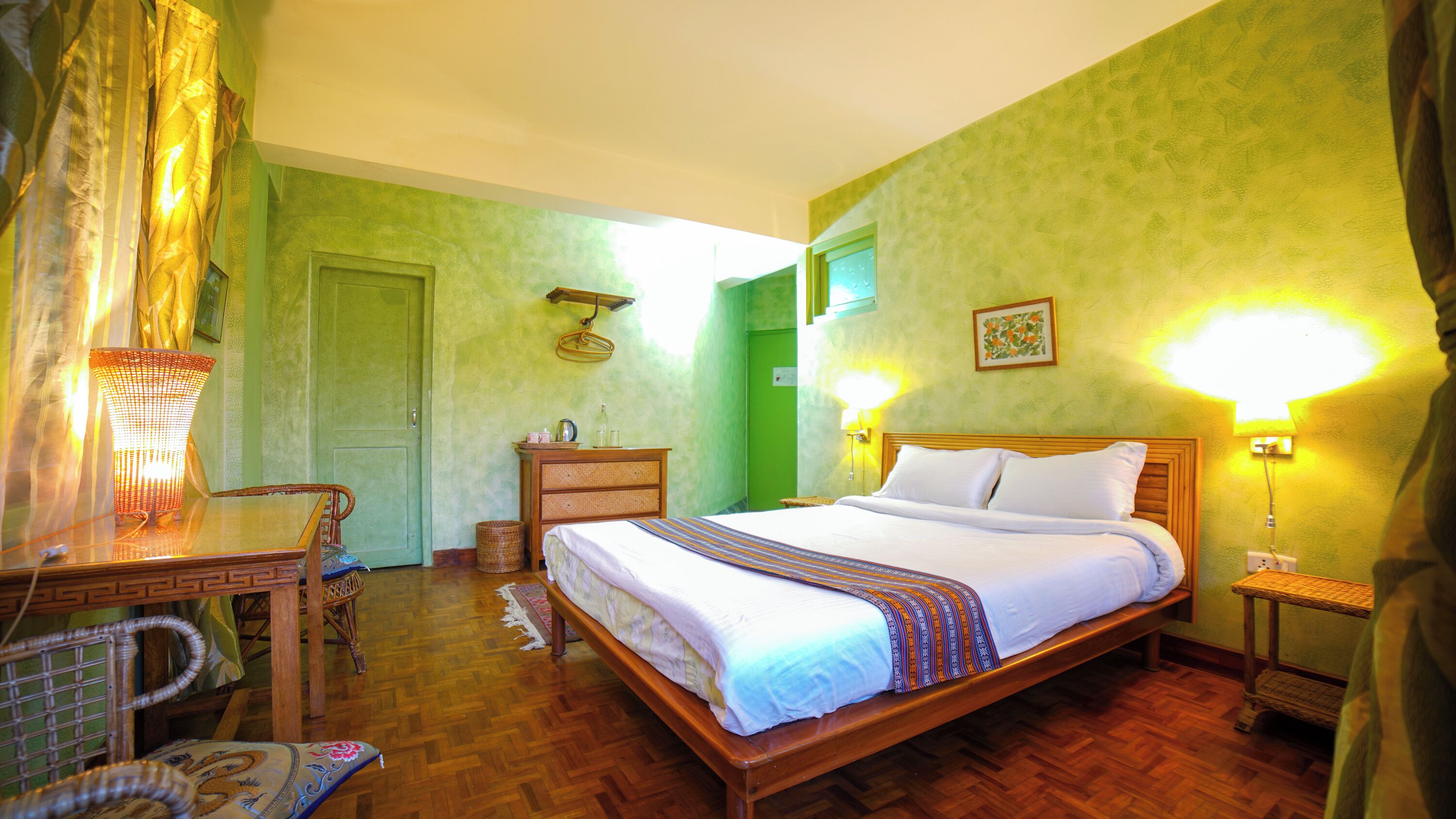 Une chambre d’hôtel avec un lit en bois, un bureau, une chaise, un panier en osier et une lampe fixée au mur.