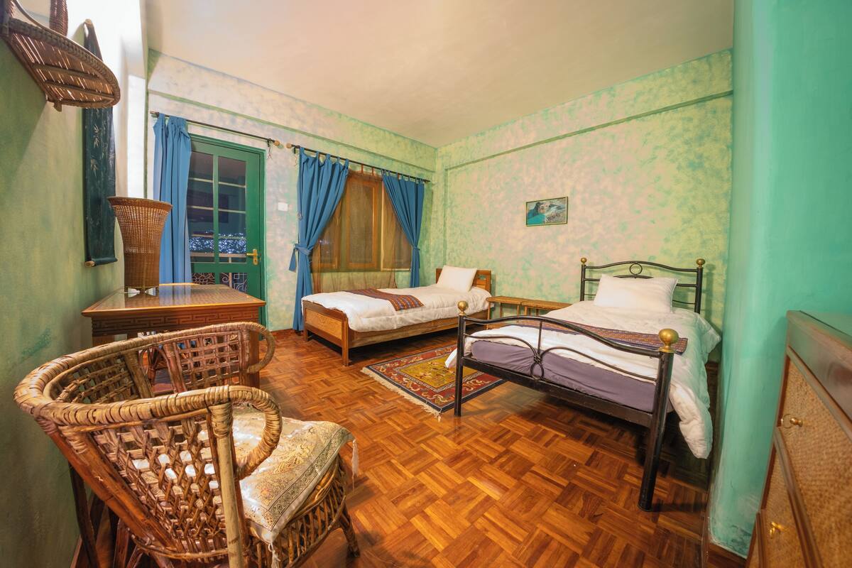 A room with two beds, a wicker chair, a wooden table, and a window with curtains.