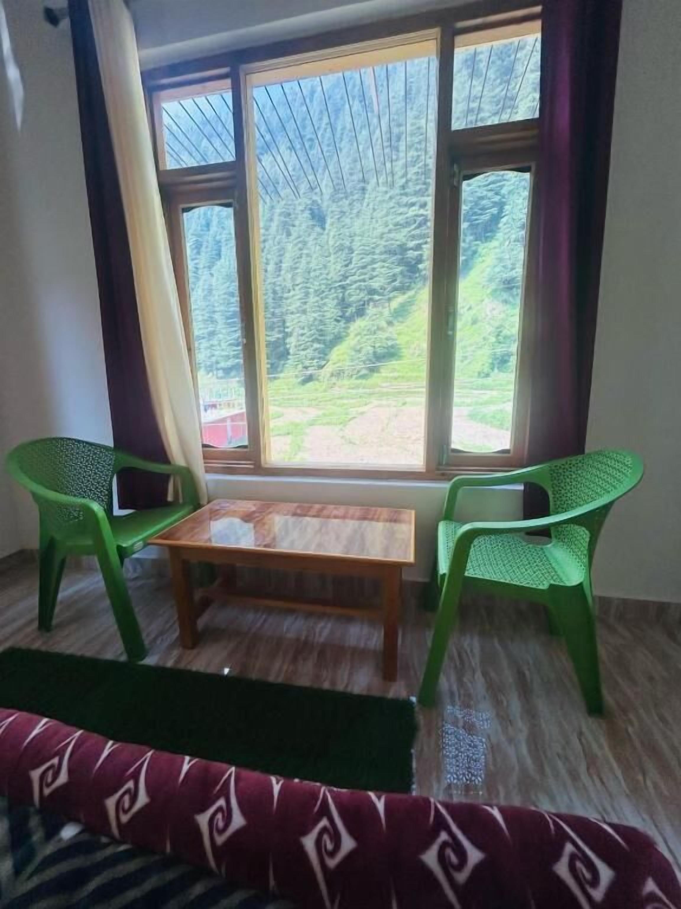 Une pièce avec une grande fenêtre, deux chaises en plastique vertes, une table en bois et un canapé à motifs.