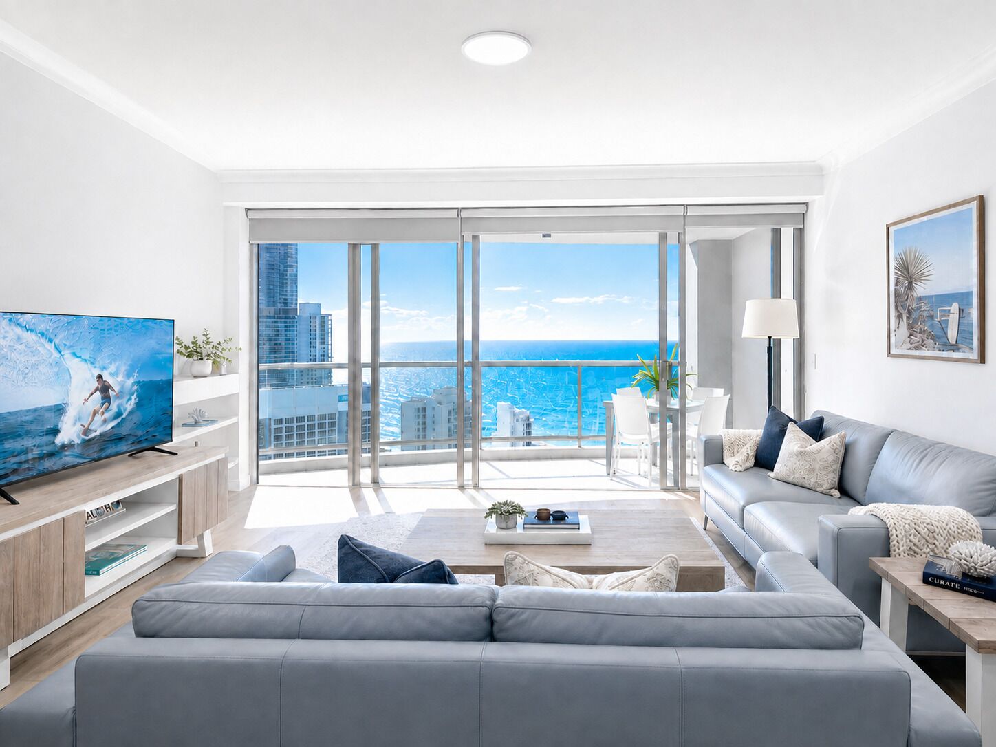 Ein modernes Wohnzimmer mit großem Fernseher, einem grauen Sofa, einem Couchtisch und Blick auf das Meer und die Stadt durch raumhohe Fenster.