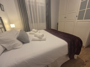 Una cama bien hecha con sábanas blancas, un almohadón gris y una manta marrón oscuro, además de dos toallas enrolladas sobre la cama.