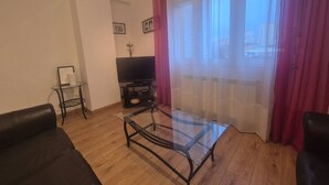 En stue med et sofabord i glas, en sort sofa, et lille sofabord, et fjernsyn og et vindue med gardiner.
