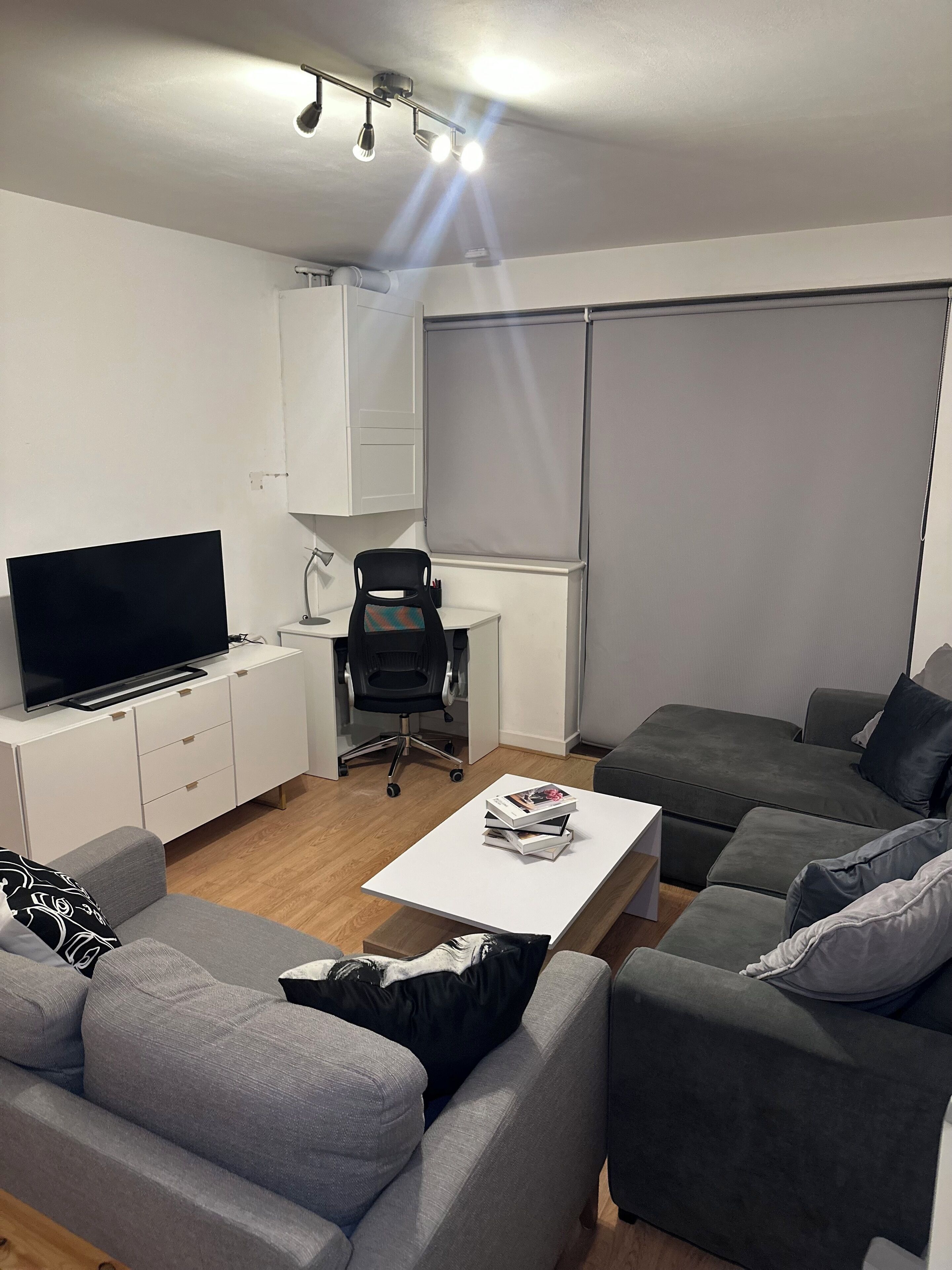 Un salon moderne avec un téléviseur à écran plat, une table basse blanche avec des livres, un canapé gris, un fauteuil gris et une grande fenêtre avec des stores.