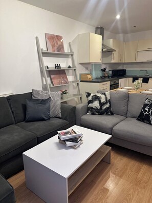 Un salón moderno con un sofá gris, una mesa de centro blanca y suelo de madera. Se ve una cocina en el fondo con armarios blancos y un microondas.