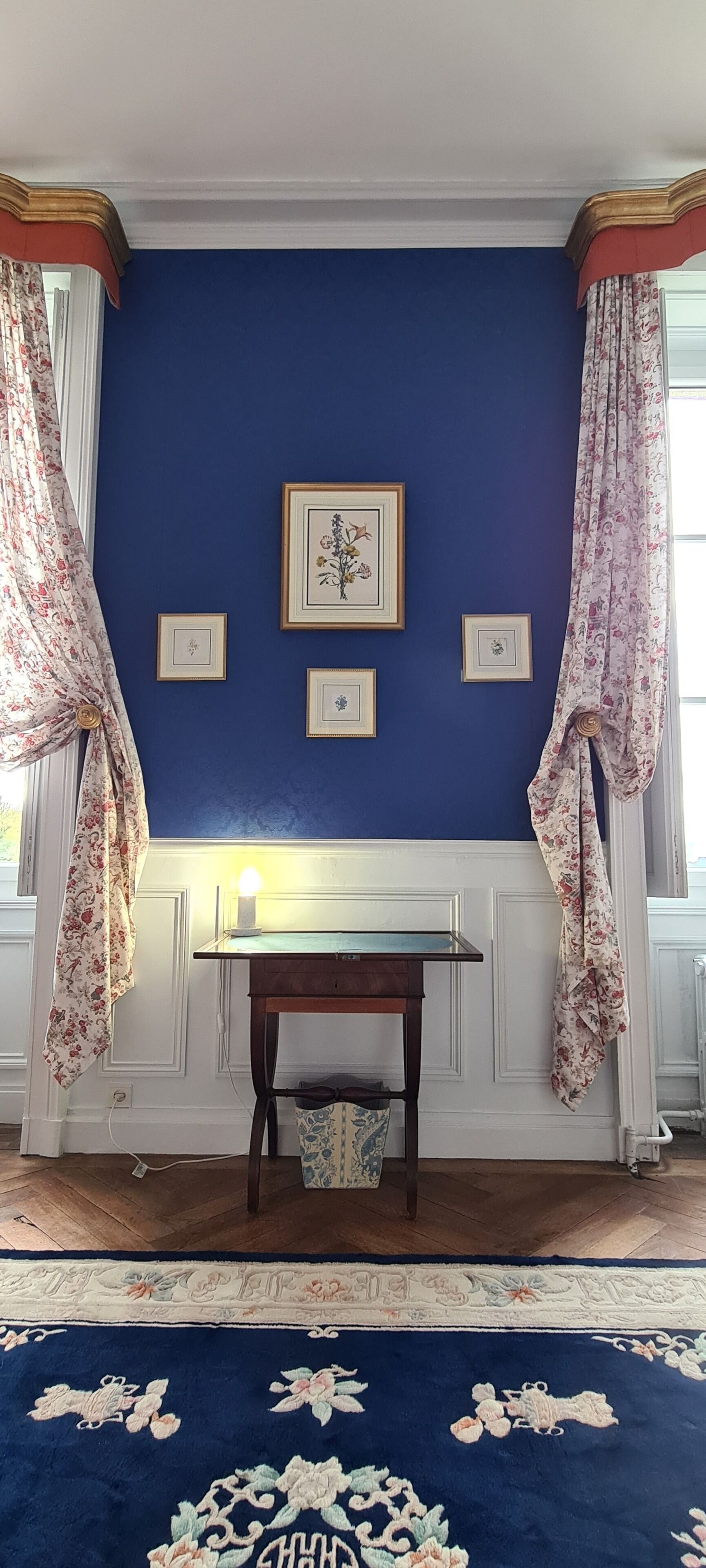 Ein Zimmer mit blauer Wand, einem Holztisch mit Lampe und blumigen Vorhängen.