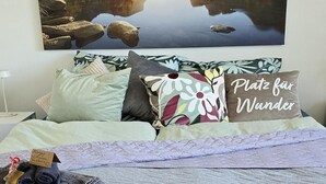 Una cama bien hecha con un edredón acolchado, un almohadón con el texto 'Platz für Wunder' y una vista de una masa de agua tranquila reflejada en un cuadro colgado.