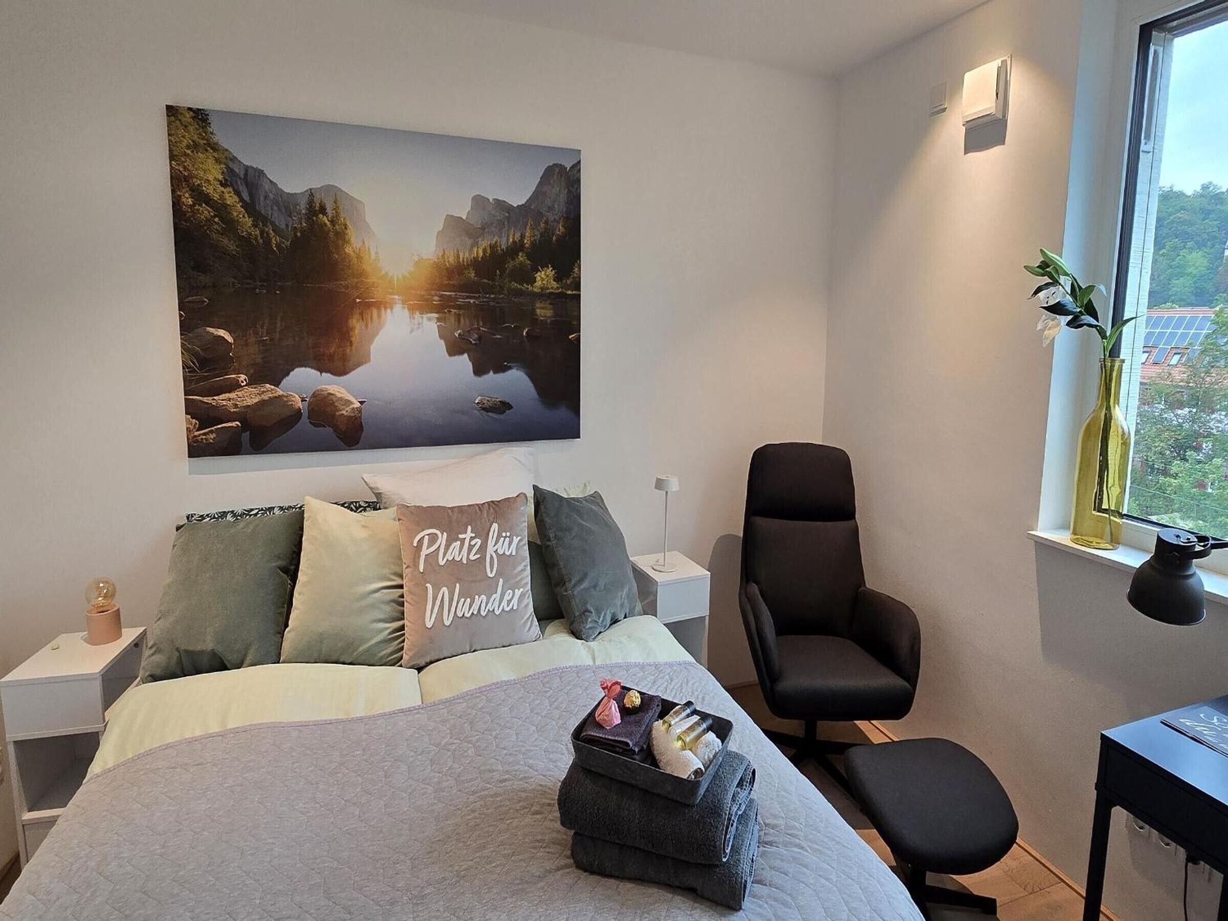 A bedroom with a bed, pillows, a chair, a bedside table, a window with a view, and a painting on the wall.