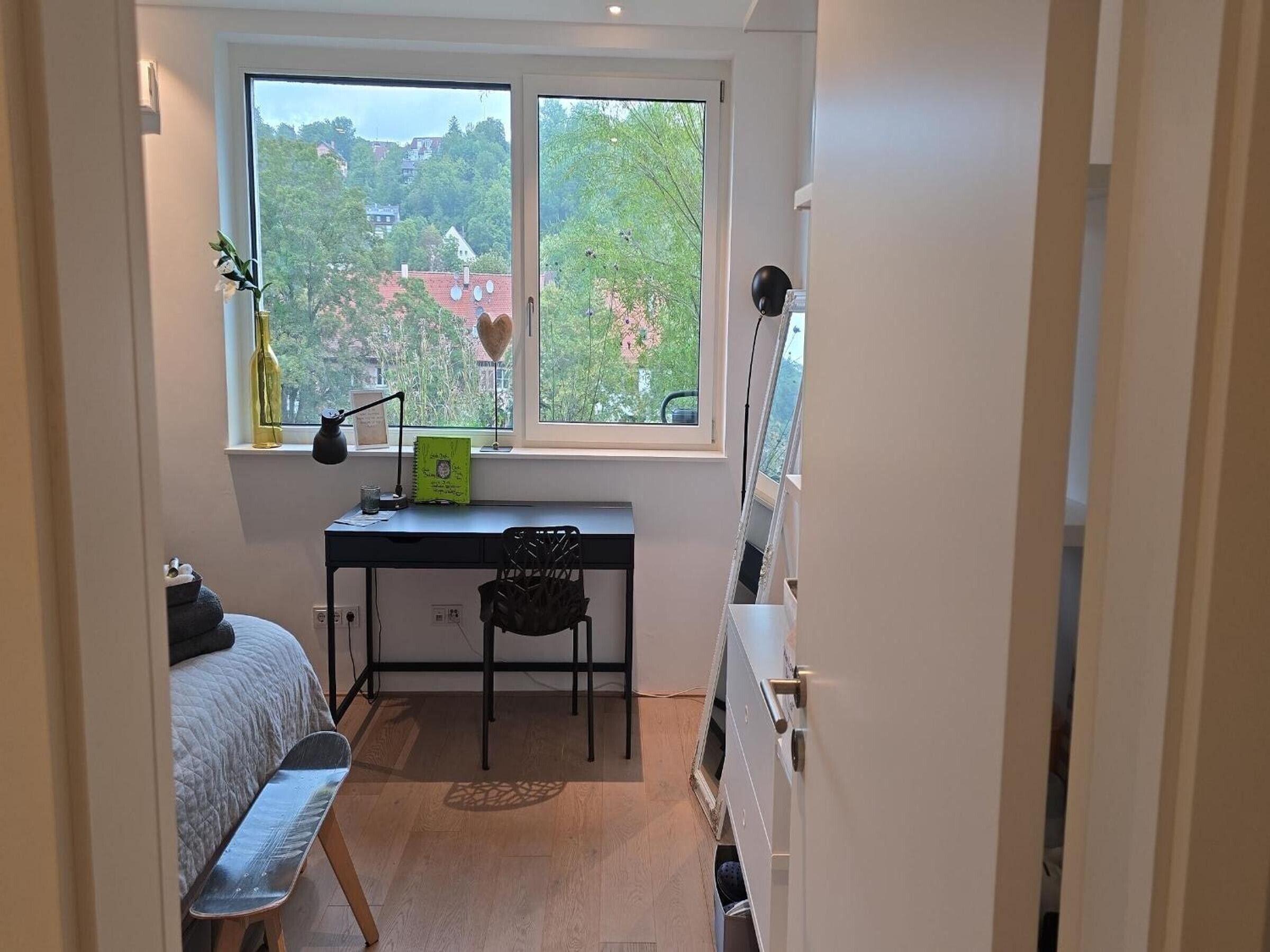 Ein kleines Schlafzimmer mit Schreibtisch, Stuhl, Bett und Fenster mit Blick auf eine grüne Landschaft und Gebäude.