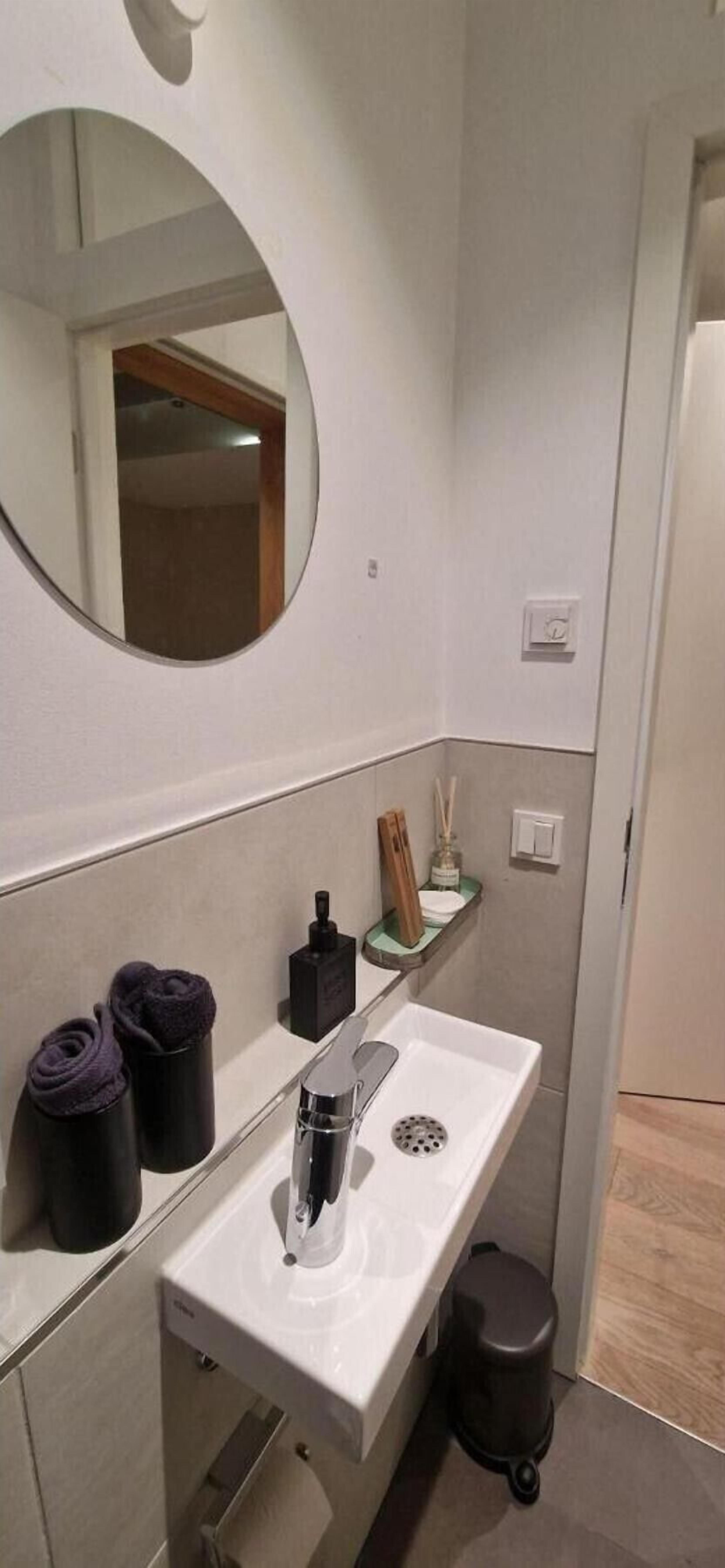Ein Badezimmer mit einem weißen Waschbecken, einem runden Spiegel, einer kleinen Ablage für Toilettenartikel und einer Mülleimer.