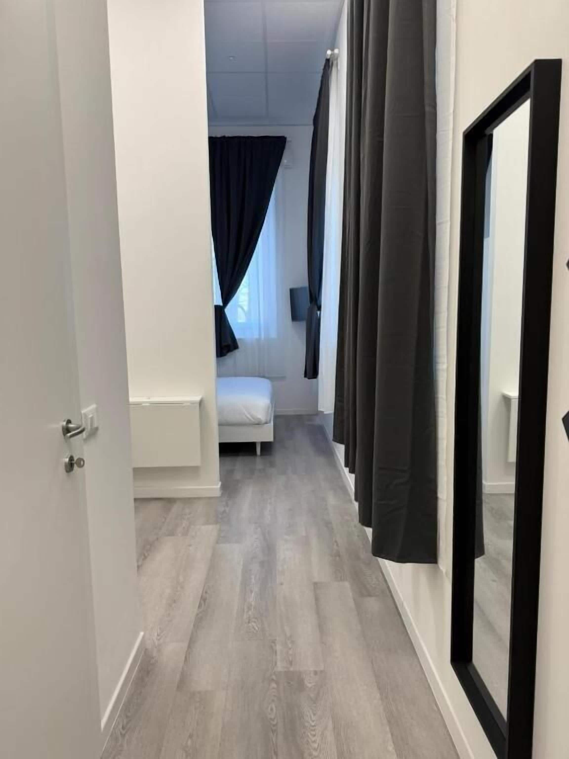 A narrow hallway with a bed and a nightstand in the background, a door on the left, and a mirror on the right.