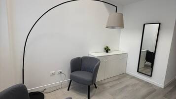 A modern room with a grey sofa, a desk with a lamp, a mirror, and a window with blinds.