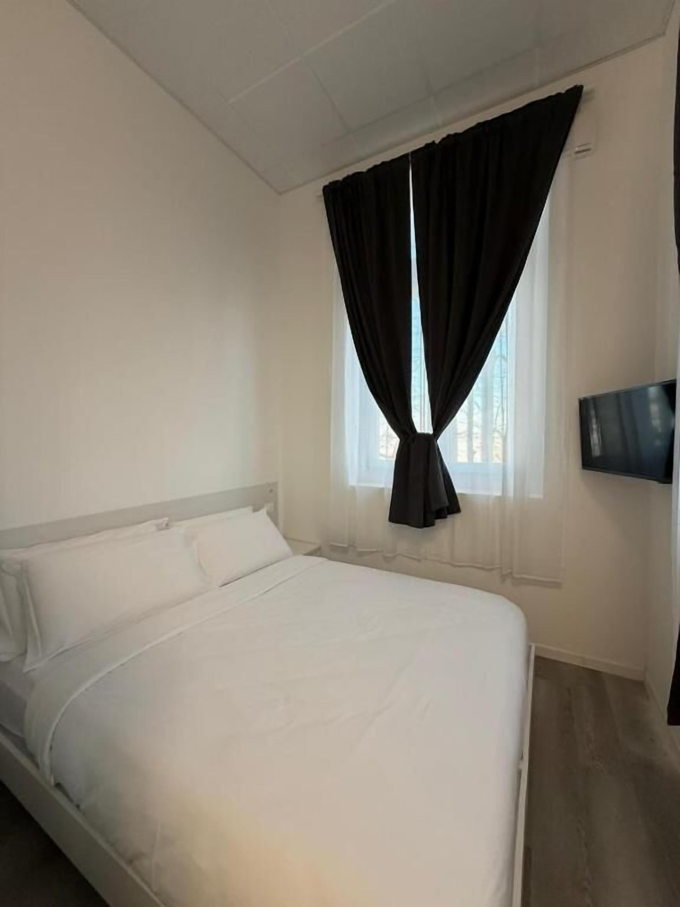 Una camera d'albergo con un letto singolo, una TV a schermo piatto fissata al muro e una finestra con tende.