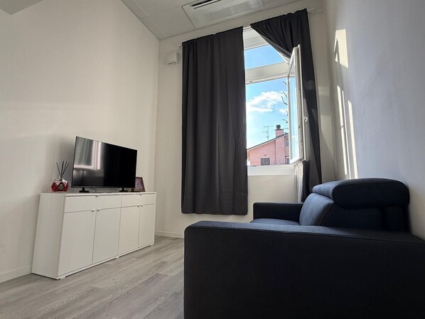 A modern living room with a flat-screen TV on a white cabinet, a dark sofa, and a window with curtains. - Viva Residence (Calderara di Reno)