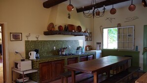 Une cuisine avec un mur recouvert de carreaux verts, des meubles en bois, une grande table en bois et des casseroles en cuivre suspendues.