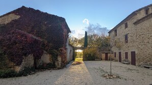 Un étroit sentier de gravier traverse une cour rustique bordée de bâtiments en pierre, dont l’un est partiellement recouvert de lierre.