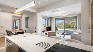 A modern kitchen with a marble countertop, a built-in oven, and a dining area with a wooden table and chairs. - Luxury carob stone villa near Almyrida beach with private pool! (Gavalohori)