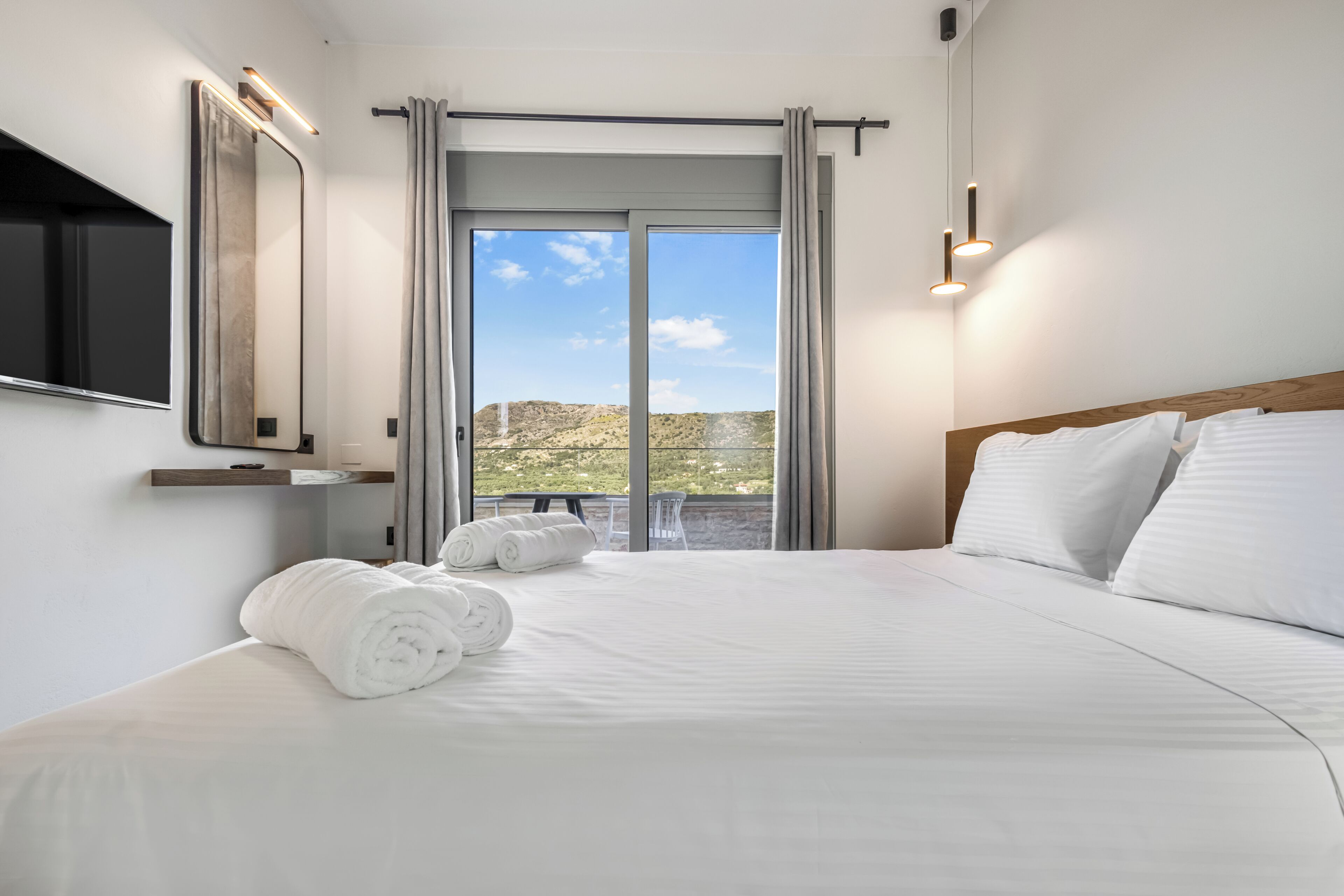 A hotel room with a bed, a TV, a balcony with curtains, and a view of the outdoors.