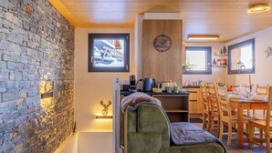 Ein gemütliches Wohnzimmer mit einer Steinwand, einem grünen Sofa, einer Essecke mit Holzkästen und einer Küchenzeile mit einer Uhr.