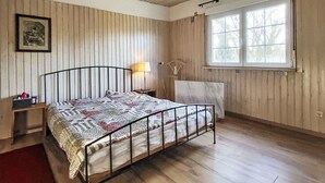 A bedroom with a wooden ceiling, a bed with a metal frame, a bedside table with a lamp, a window with curtains, and a framed picture on the wall.