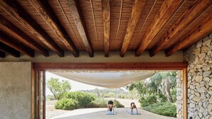 Un espacio interior con techo de madera y paredes de piedra, que conduce a una zona exterior con terraza de madera y vegetación.