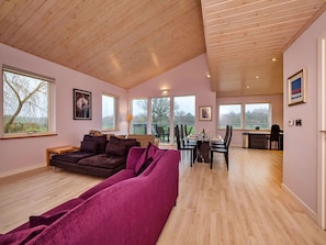 Ein großzügiger Wohnbereich mit Holzboden, einem violetten Sofa, einem Esstisch mit Stühlen und großen Fenstern mit Blick ins Freie.