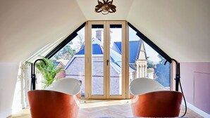 Een kamer met een schuine plafond, twee oranje stoelen, een blauw vloerkleed en uitzicht op een gebouw met een rood dak door een glazen deur.