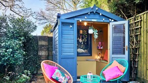 Een blauwe schuur met een geopende deur, waar een schilderij en een hanglamp zichtbaar zijn. Buiten staan twee rieten stoelen met kleurrijke kussens.