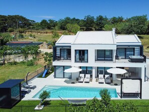 Ein modernes, zweistöckiges Haus mit Swimmingpool, Sitzgelegenheiten im Freien und Blick auf die umliegende Landschaft.