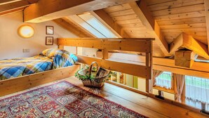 Une chambre à coucher aménagée dans un espace sous les toits, avec un plafond en pente, des poutres en bois, un lit recouvert d’une couette à carreaux, un tapis à motifs et un panier rempli de plantes vertes.