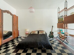 Ein Zimmer mit Schachbrettfußboden, einem Bett mit dunkler Decke, einer Gitarre, einer Lampe und einem Spiegel.