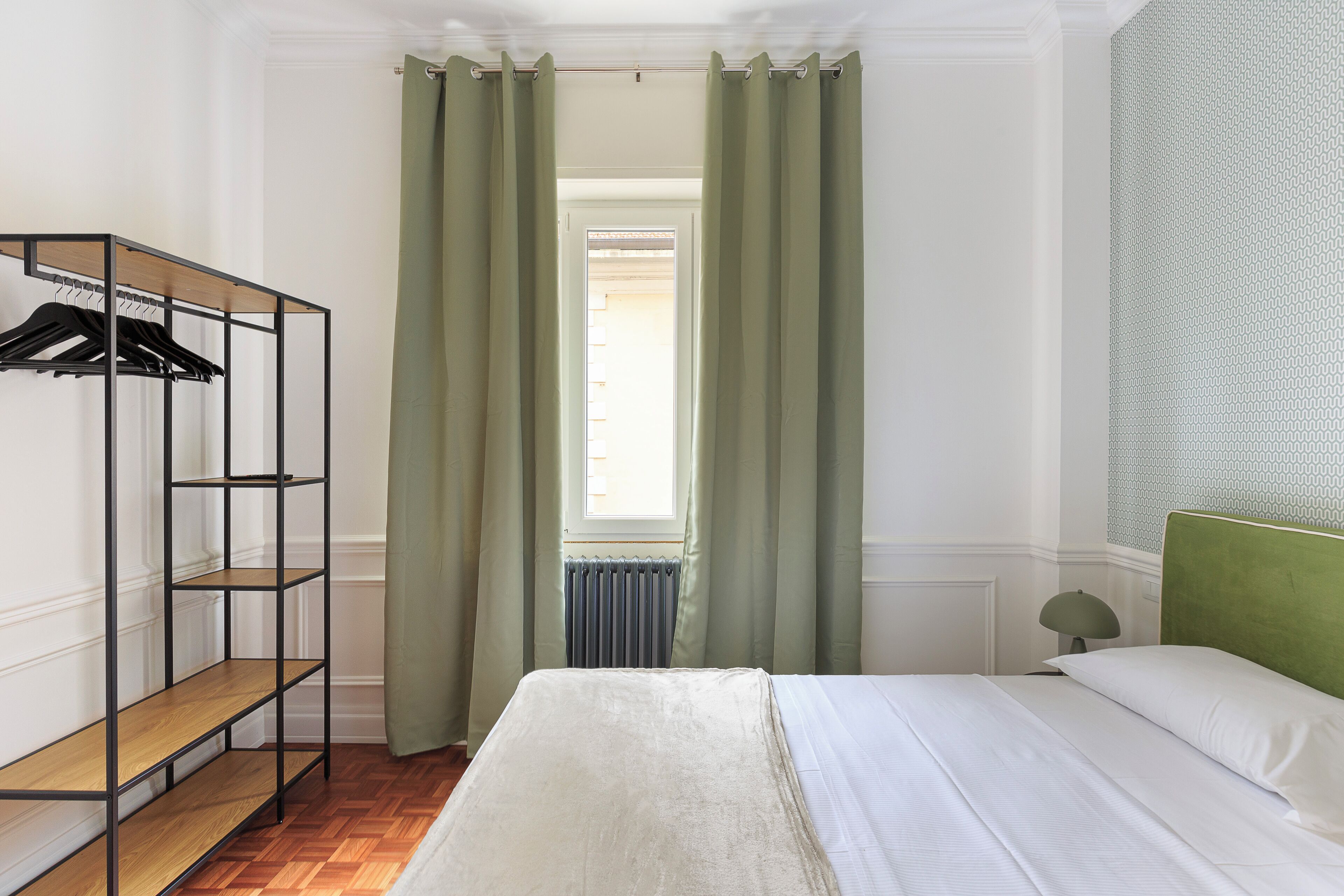 Ein Schlafzimmer mit einem Bett, einer Holzregal-Einheit mit Kleiderhaken, einem Heizkörper und einem Fenster mit Vorhängen.