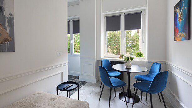 Un coin repas comprenant une table ronde, quatre chaises bleues, une petite table d’appoint et une fenêtre donnant sur un paysage verdoyant.
