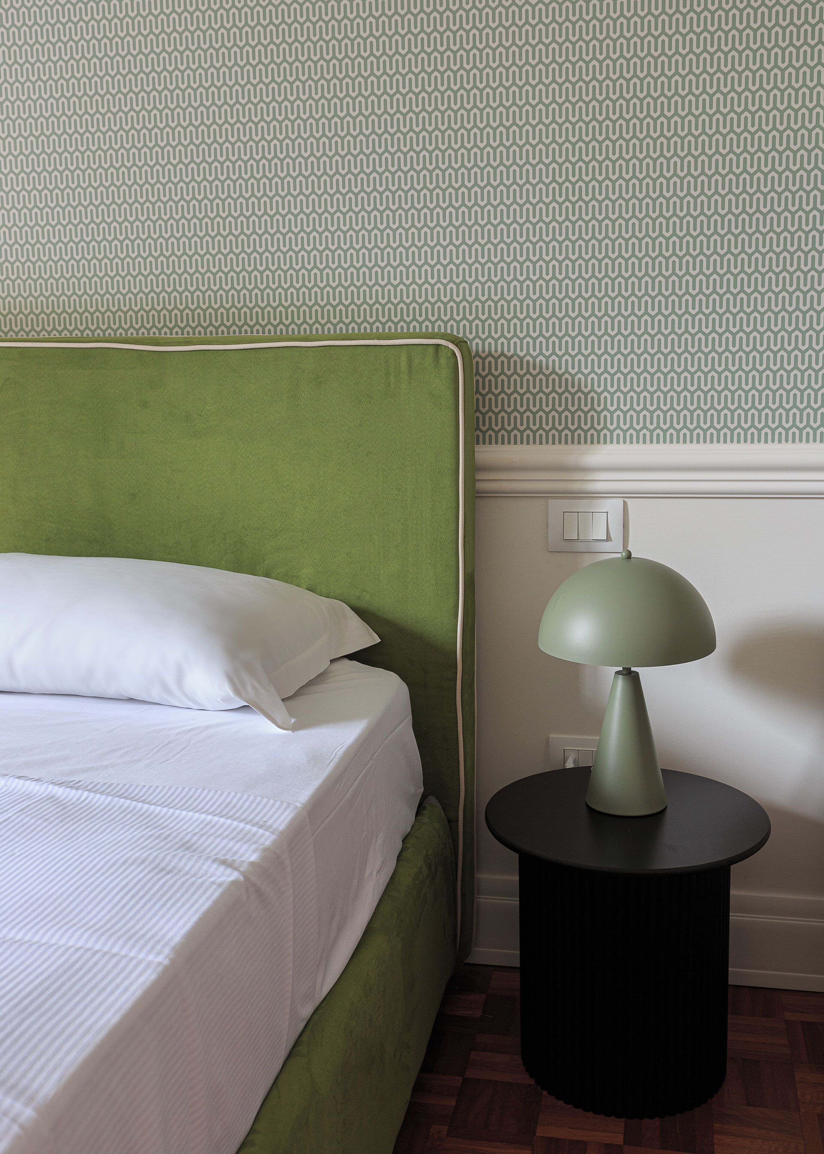 A bed with white bedding, a green headboard, and a black side table with a lamp.