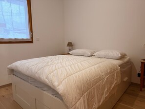 Una cama individual con edredón blanco acolchado, una mesita de noche con lámpara y una ventana con vistas a árboles.