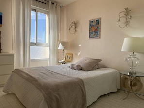 A bedroom with a bed, a window with curtains, a bedside table with a lamp, and wall art. - Cozy T3 Apartment with Terrace in Canet-en-Roussillon (Canet-en-Roussillon)