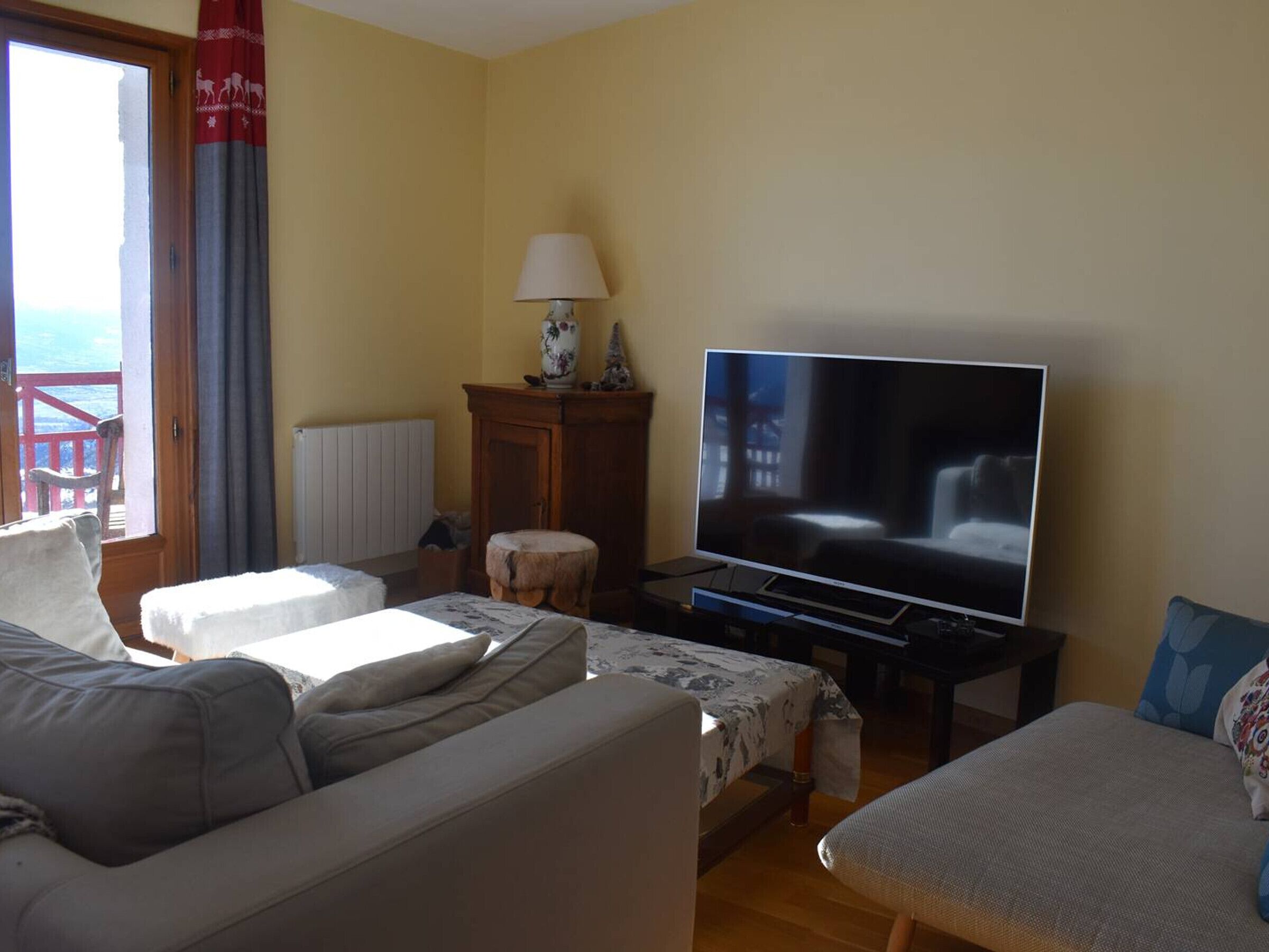 A living room with a large flat-screen TV, a wooden cabinet, a lamp, and a view of the sea through a window.