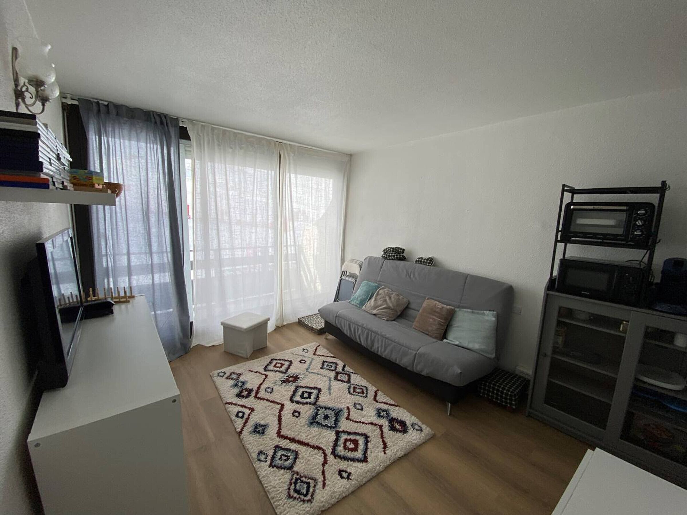 Un espace de vie compact avec un canapé gris, un tapis à motifs et un téléviseur posé sur un meuble.