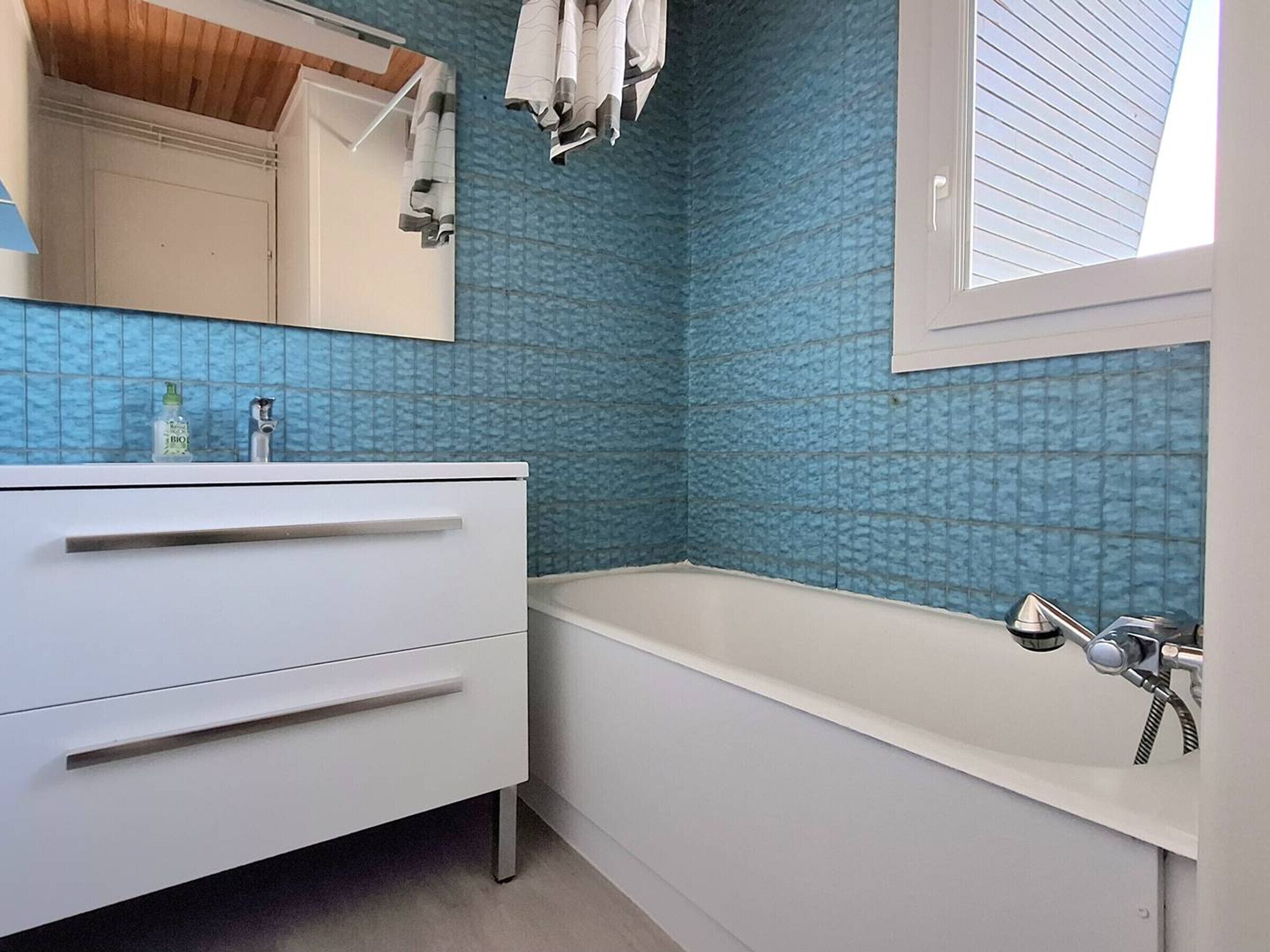 Un baño con tocador blanco, pared con azulejos azules, una ventana y una bañera.