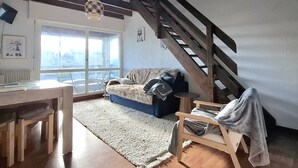 A cozy living room with a wooden staircase, a sofa, a small table, and a view through a large window. - Cozy T2 Cabin with enclosed loggia (Bolquère-Pyrénées 2000)