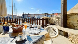 Ein Tisch im Freien mit Frühstücksangeboten, Blick auf Boote und Gebäude sowie eine Steinmauer.