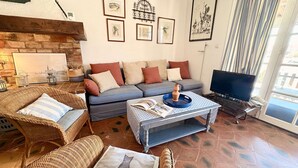 Ein Wohnzimmer mit einer Couch, zwei Korbstühlen, einem Couchtisch mit Büchern, einem Fernseher und Kunstwerken an den Wänden.