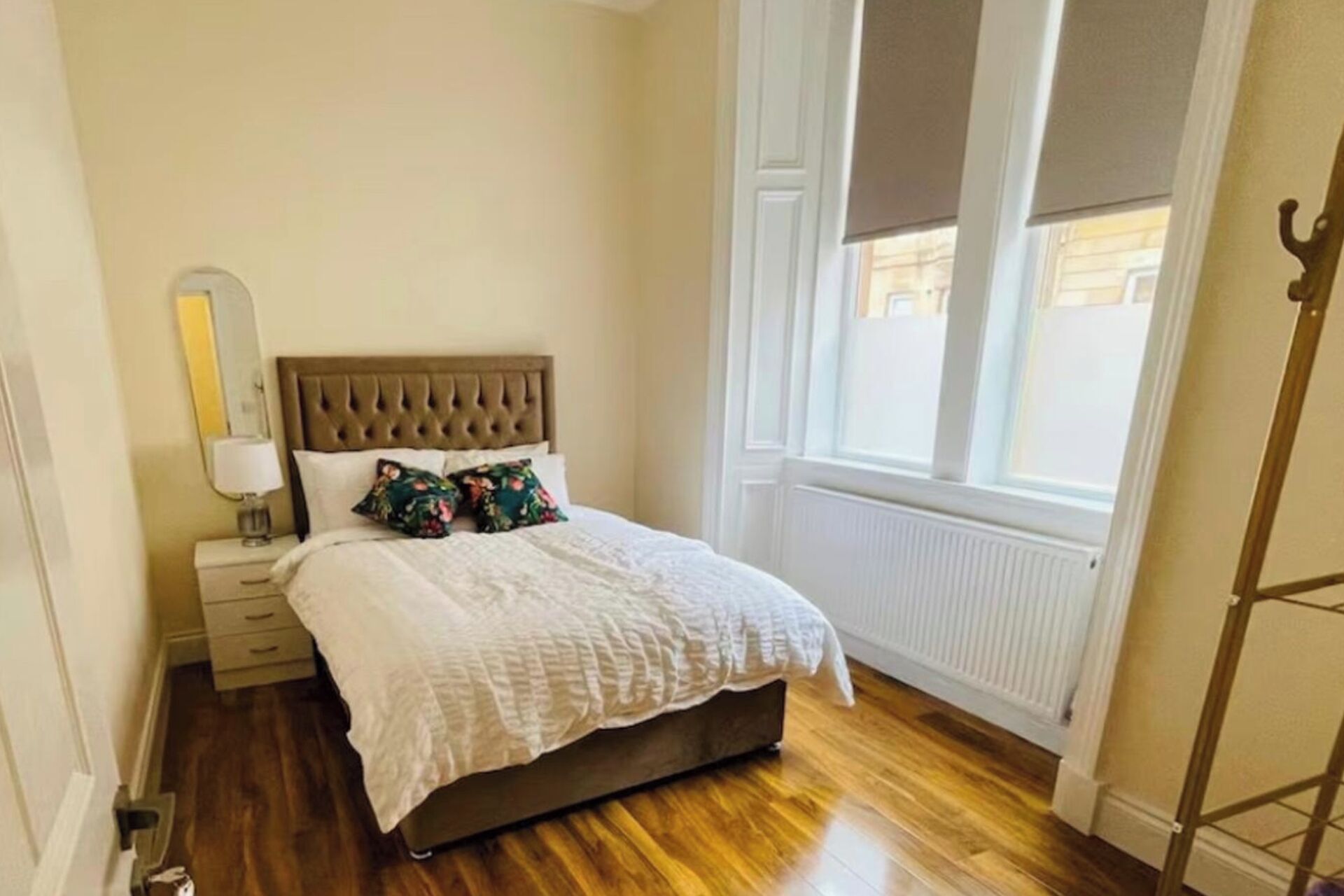Een slaapkamer met een houten vloer, een bed met een witte dekbedovertrek en bloemenkussens, een nachtkastje met een lamp en een raam met jaloezieën.