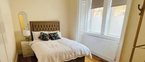 Ein Schlafzimmer mit Holzboden, einem Bett mit weißer Bettdecke und floralen Kissen, einem Nachttisch mit Lampe sowie einem Fenster mit Jalousien.