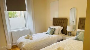 A hotel room with two beds, each with a headboard, a bedside table with a lamp, and a window with blinds. - Near Hampden - Parking - Wifi - Sleeps 6 (Glasgow)