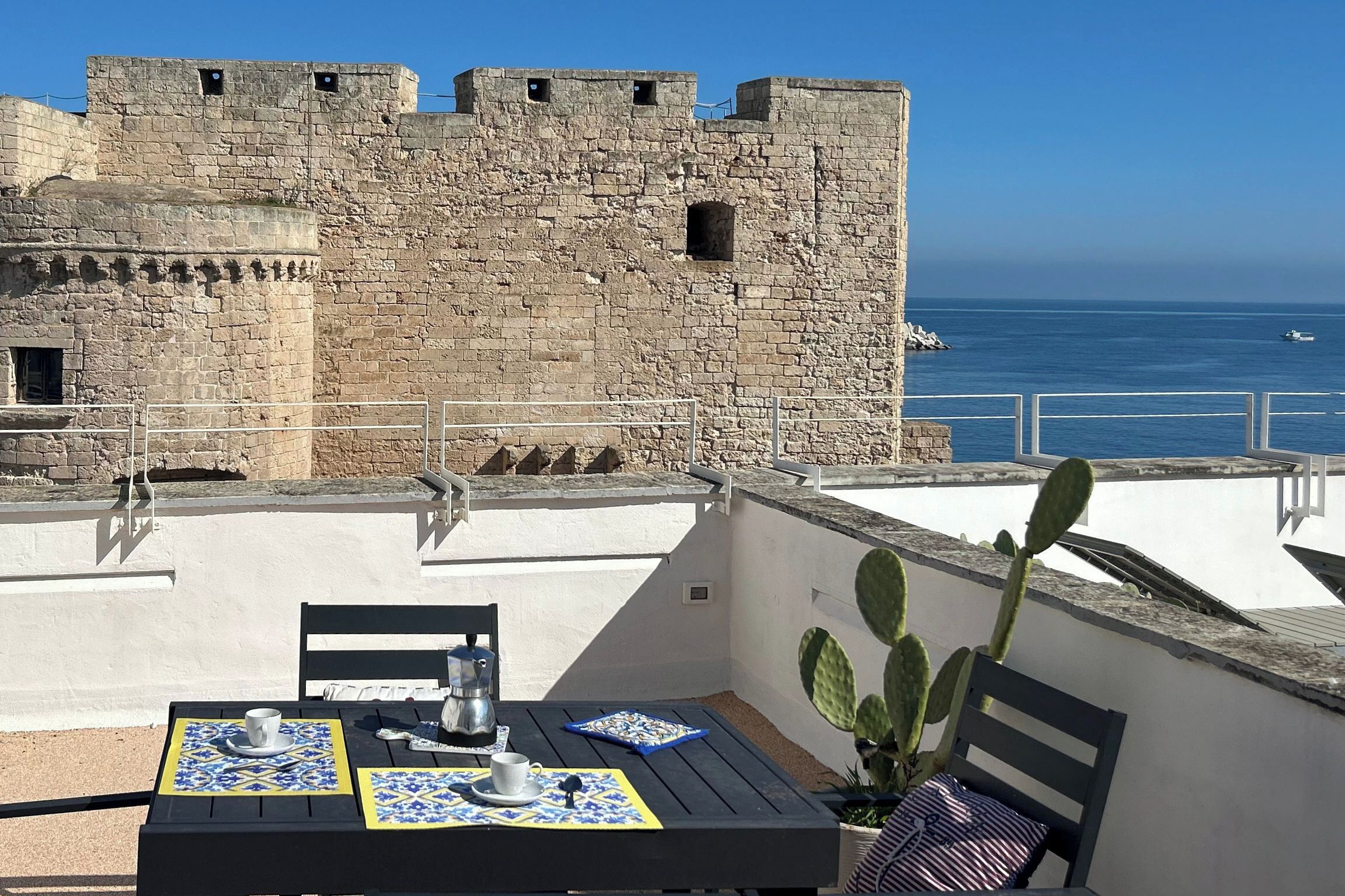 Terraço com mesa posta para dois, com vista para uma muralha de pedra histórica e o mar.