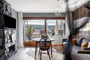 Ein Wohnzimmer mit Kamin aus Stein, einem Flachbildfernseher, einem runden Esstisch mit einem Stuhl, einer Couch und einem großen Fenster mit Blick auf einen bewaldeten Hügel.