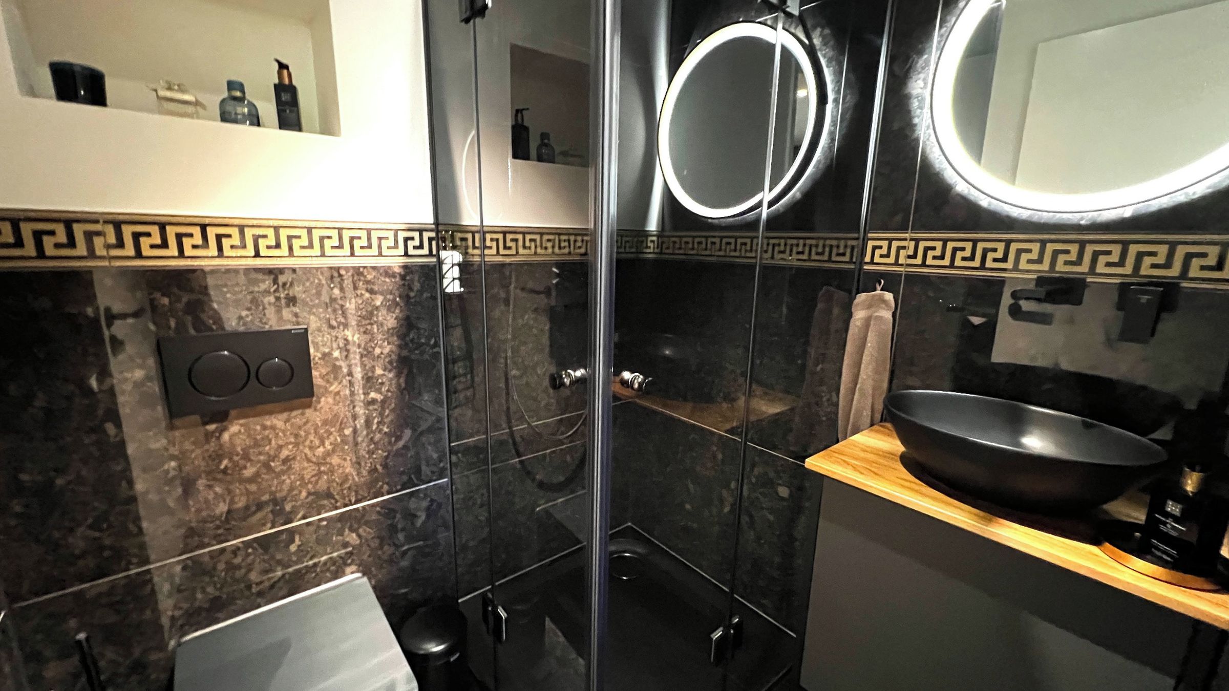 A modern bathroom with a black marble wall, a wooden vanity, a black sink, and a mirror with a circular frame.