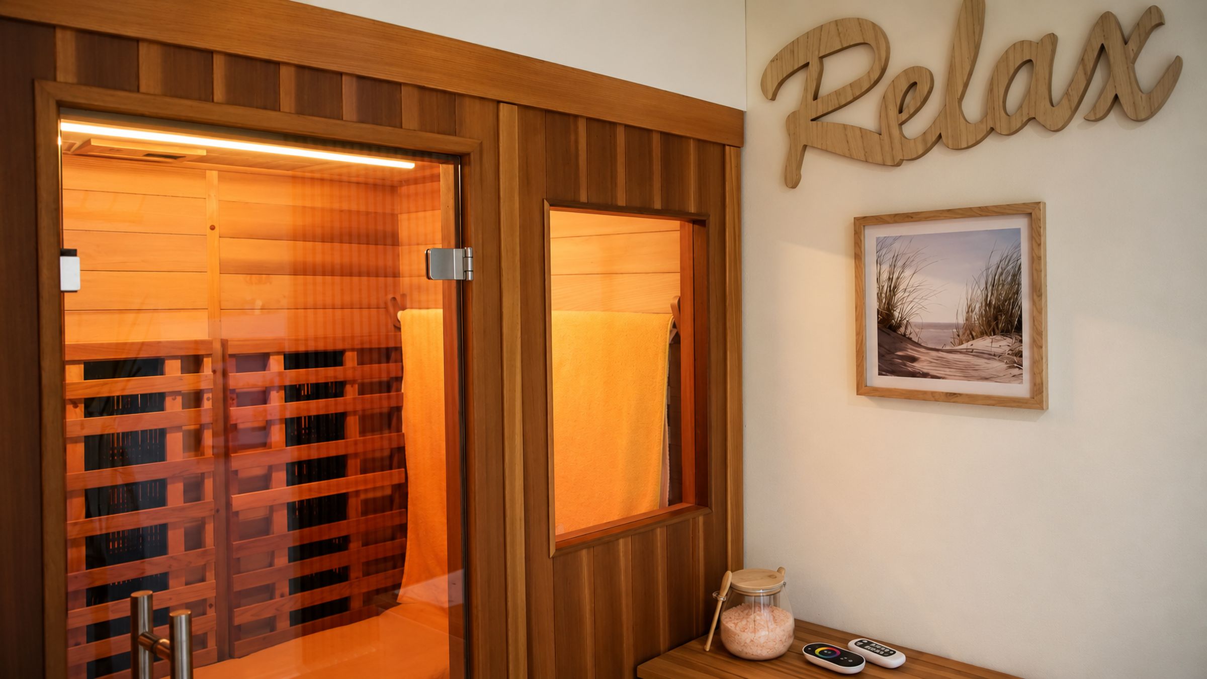 A wooden sauna with a sliding door, a framed picture on the wall, and a small table with a jar and remote controls.