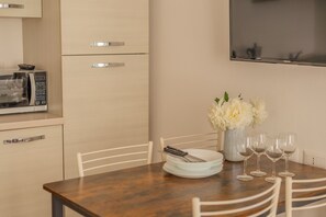 Een eethoek met een houten tafel, witte stoelen, een witte vaas met bloemen en een flatscreen televisie aan de muur.